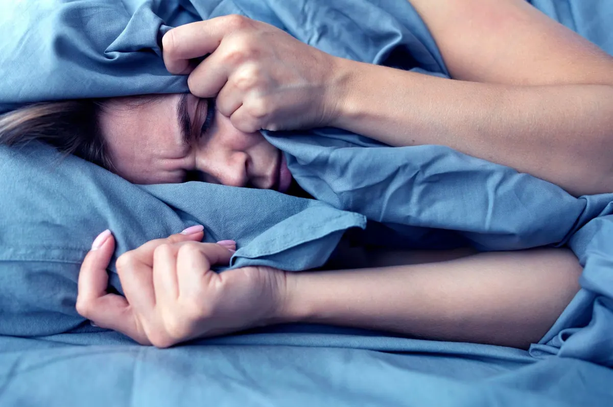A woman in bed pulling the sheets over her head, unable to sleep
