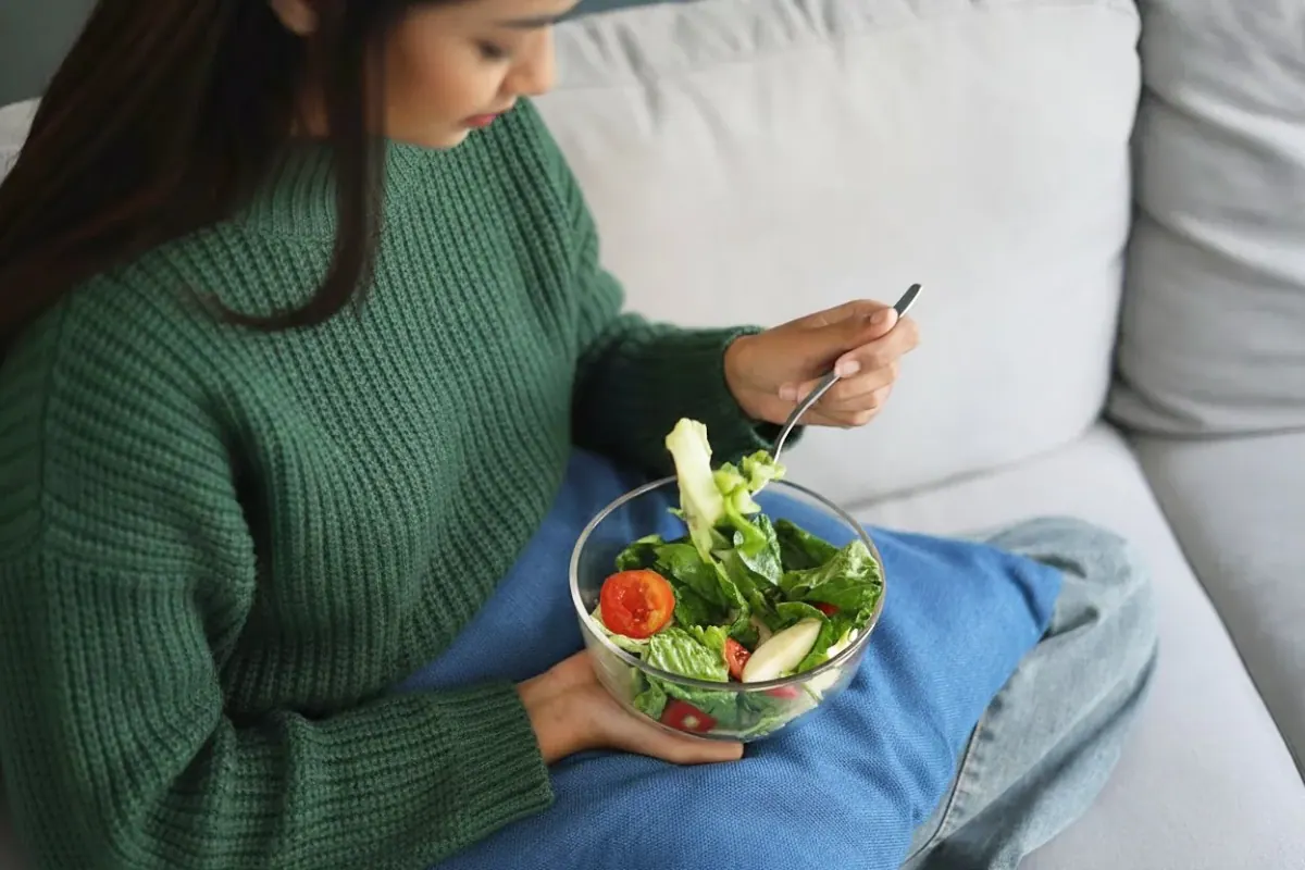 A smiling woman in green holding a vibrant whole-food bowl with fresh vegetables on the table