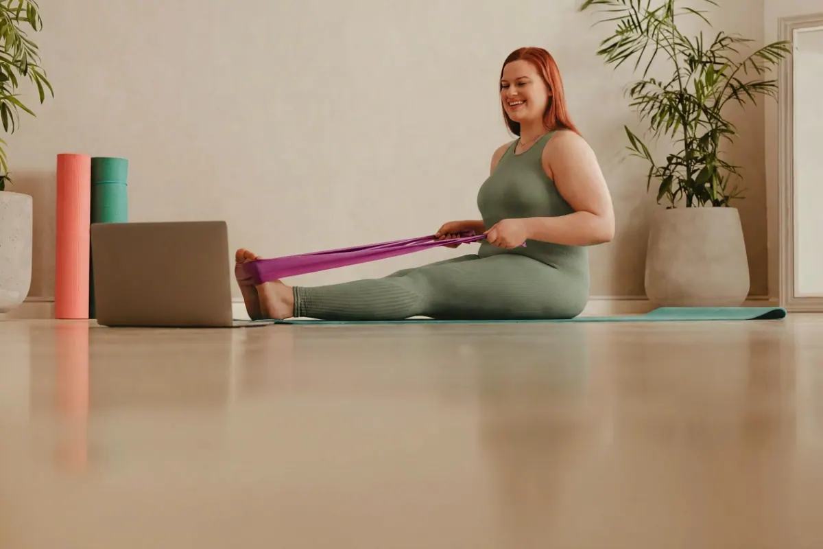 A smiling woman in athletic wear doing a guided resistance-band workout on a mat at home in front of a laptop